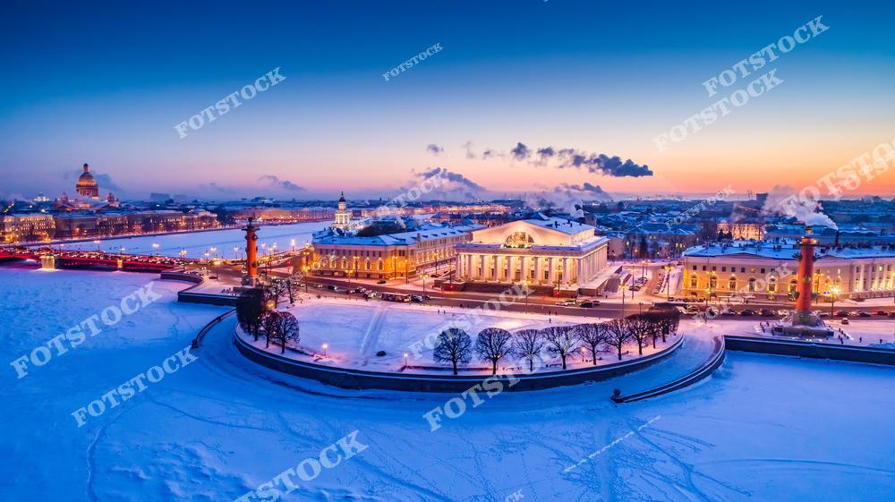 На фото изображена Стрелка Васильевского острова, обрамлённая замёрзшей Невой. Здание Биржи, ныне музей, и Ростральные колонны выделяются на фоне вечернего неба, которое окрашено в мягкие зимние оттенки заката. Вдали можно увидеть Исаакиевский собор, дополняющий классический городской пейзаж. Этот вид подчёркивает красоту и величие зимнего Петербурга в вечерние часы.